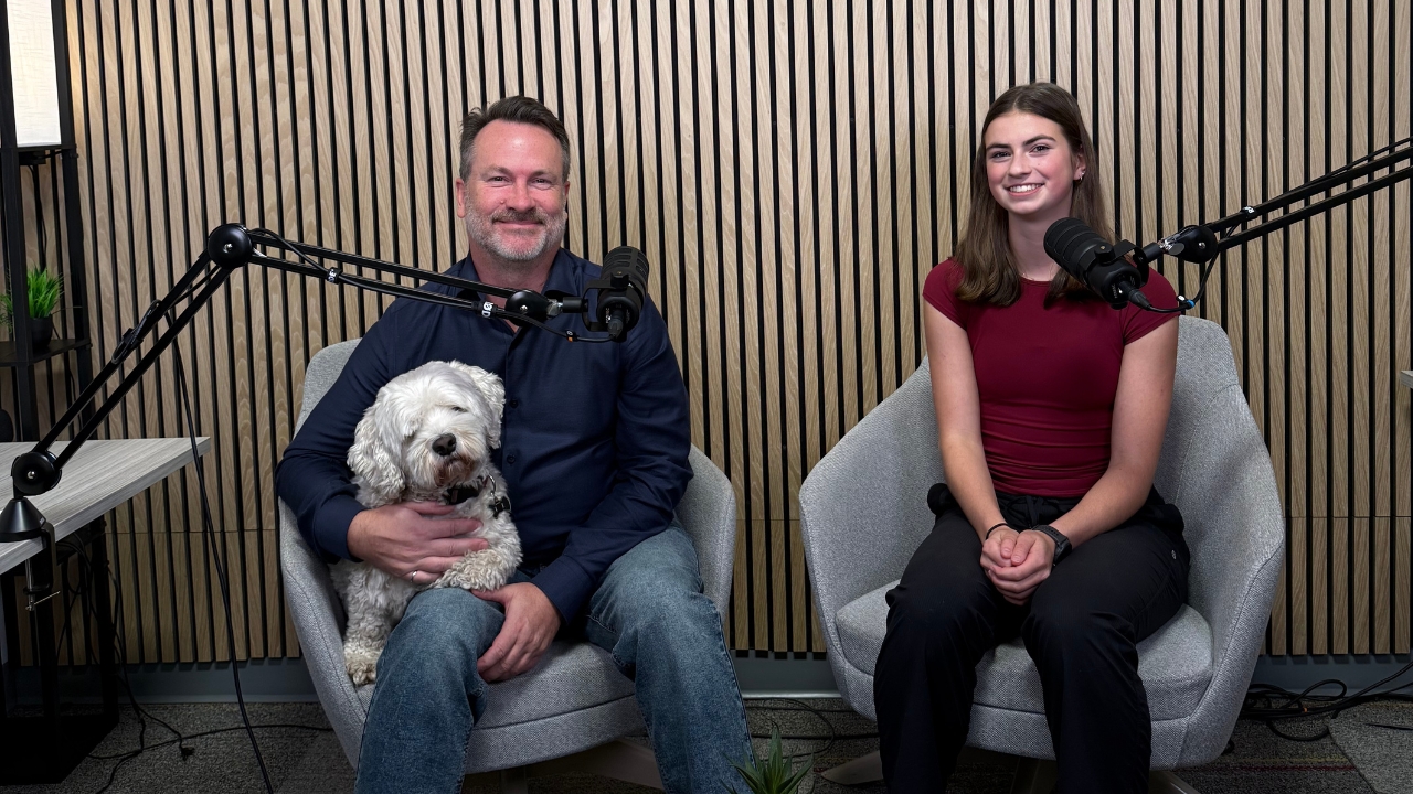 A man with a white dog on his lap sits next to a young woman in a podcast booth